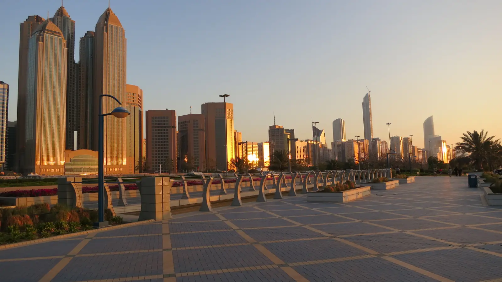 Calçadão da Corniche de Abu Dhabi ao pôr do sol, com canteiros, postes de luz e os arranha-céus da orla iluminados pela luz dourada.