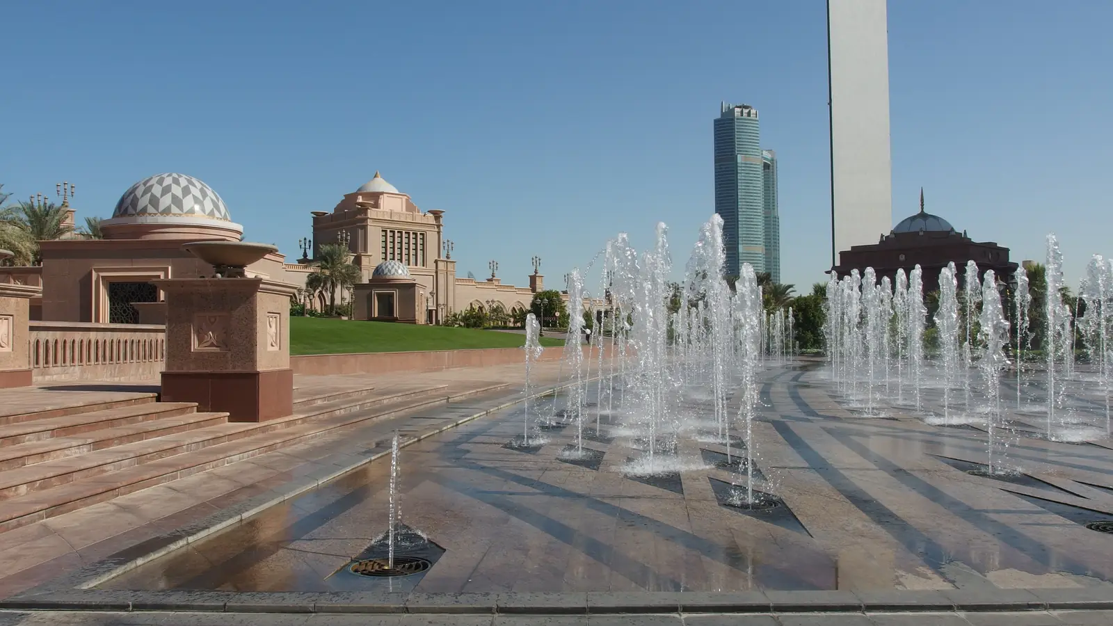 Fontes na entrada do Emirates Palace, com as cúpulas e a fachada cor de areia do hotel e torres modernas ao fundo, sob céu azul.