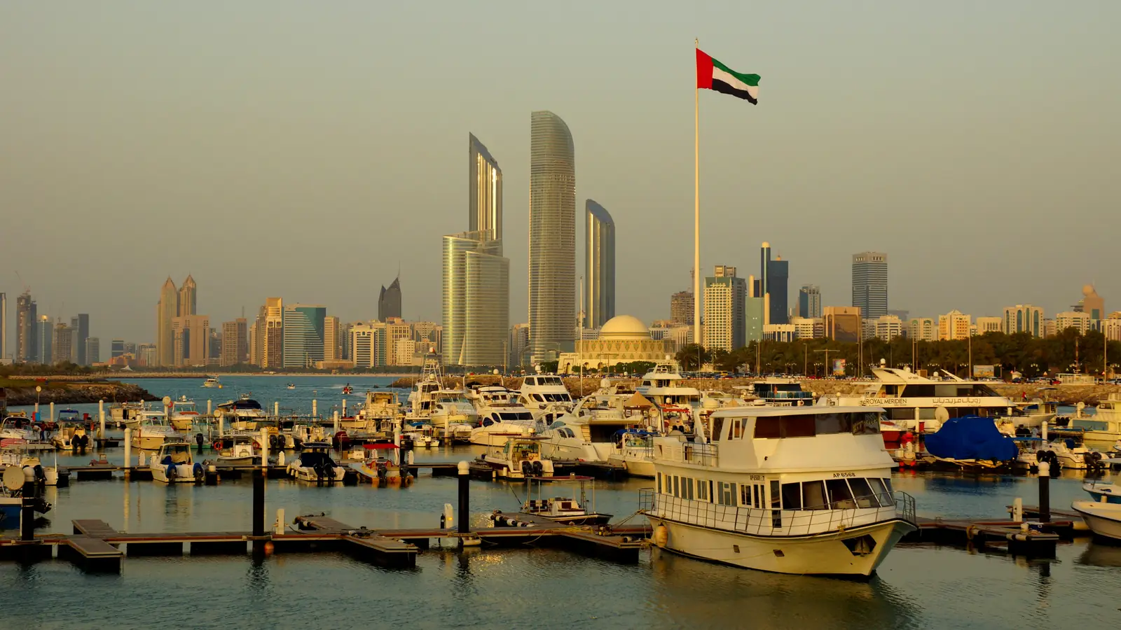 Marina de Abu Dhabi ao entardecer, com iates atracados, a bandeira dos Emirados e os arranha-céus da cidade ao fundo.