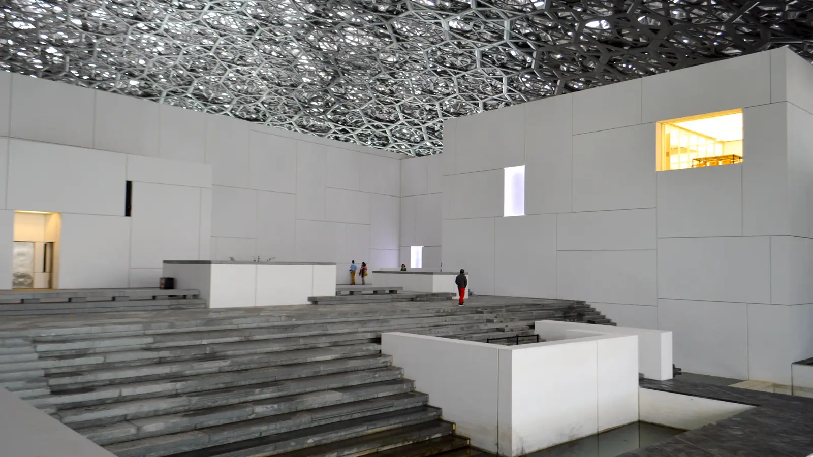 Interior do Louvre Abu Dhabi: volumes brancos, escadarias e visitantes sob a cúpula rendada de metal que filtra a luz.