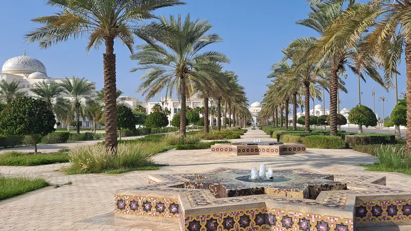 Jardins do Qasr Al Watan com fonte de mosaico, alameda de palmeiras e as cúpulas brancas do palácio ao fundo, sob céu azul.