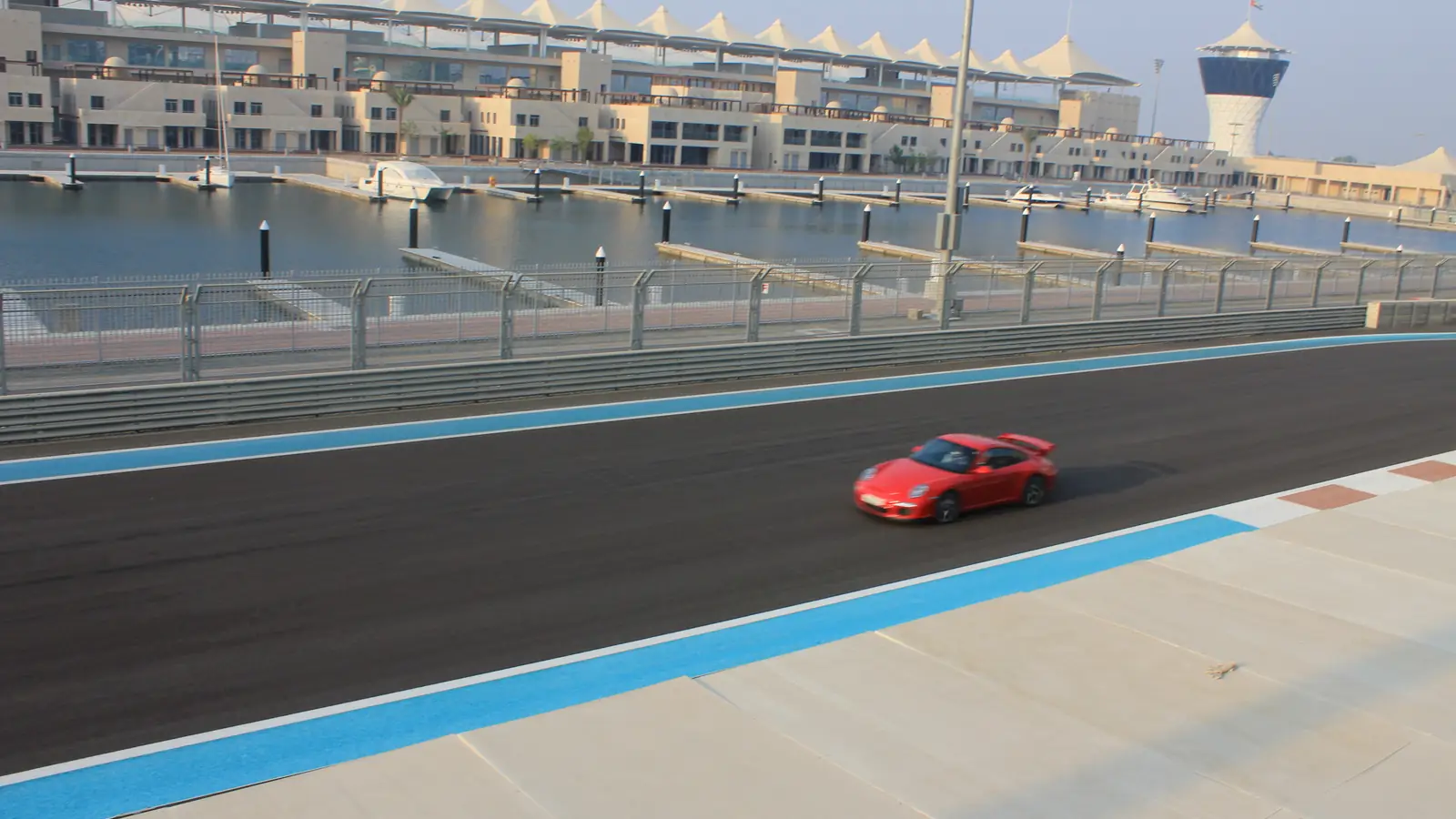 Carro esportivo vermelho na pista do circuito Yas Marina, com a marina, iates e a torre de controle ao fundo.