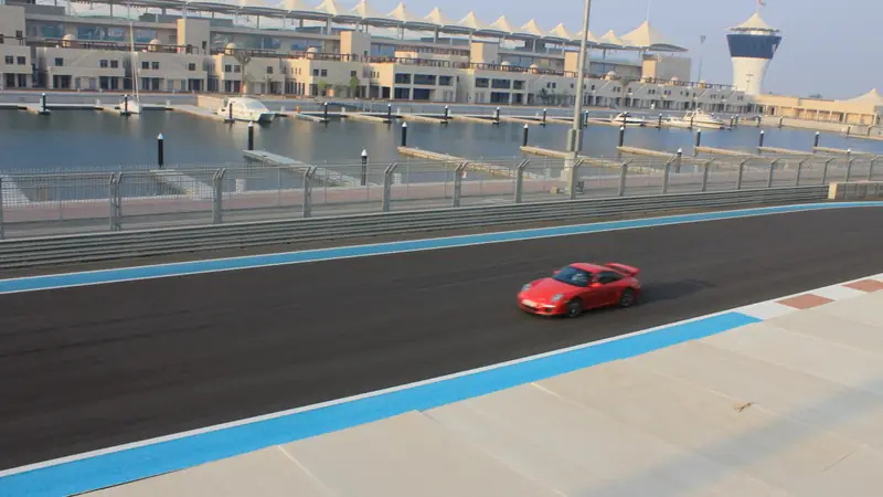 Carro esportivo vermelho na pista do circuito Yas Marina, com a marina, iates e a torre de controle ao fundo.