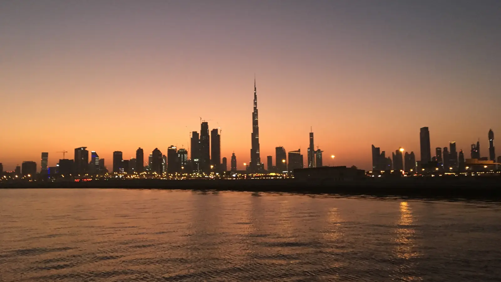Skyline de Dubai ao entardecer, com o Burj Khalifa acima das torres e as luzes refletidas na água.