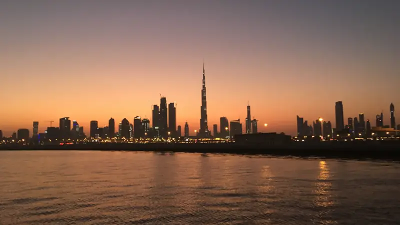Skyline de Dubai ao entardecer visto do outro lado da água, com a silhueta do Burj Khalifa acima das torres, céu alaranjado e luzes refletidas.