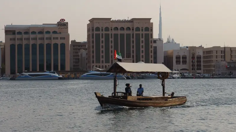 Abra de madeira com toldo e bandeira dos Emirados cruzando o Dubai Creek, com prédios da orla atrás e o Burj Khalifa ao fundo, à distância.