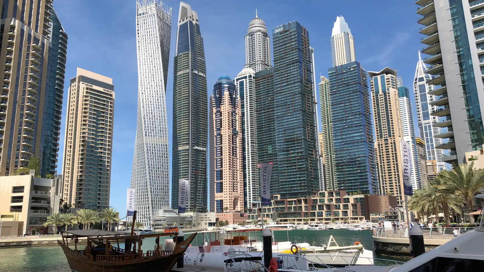 Dubai Marina: torres altas, entre elas um arranha-céu retorcido, erguem-se sobre a água, com um dhow de madeira e barcos brancos atracados em primeiro plano.