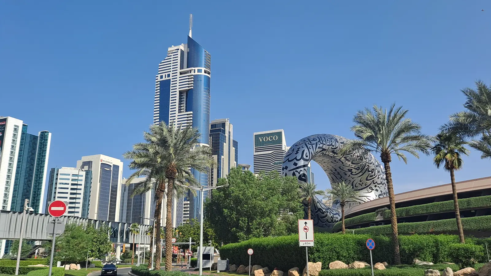 O Museu do Futuro, em forma de anel prateado coberto de caligrafia árabe, entre palmeiras e torres de vidro da Sheikh Zayed Road, sob céu azul.