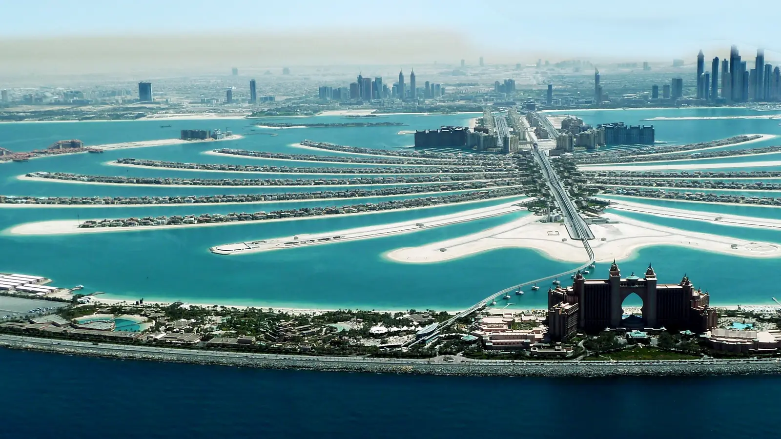 Vista aérea da Palm Jumeirah: as folhas da palmeira e o crescente em água turquesa, o hotel Atlantis em primeiro plano e as torres da Dubai Marina no horizonte.